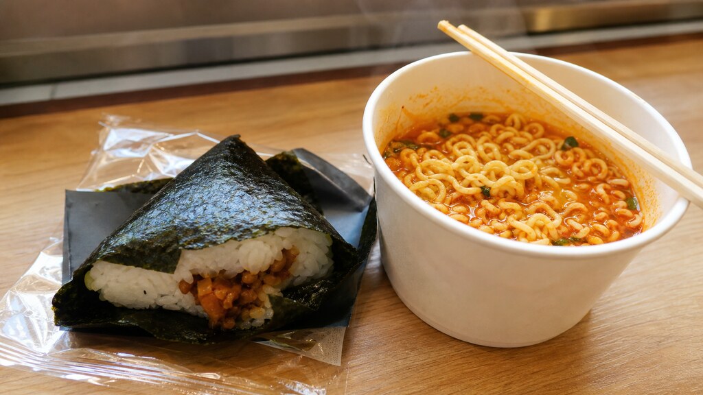 A triangular nori-wrapped rice ball partly peeled open next to a steaming cup of instant ramen with wooden chopsticks resting on a convenience-store eat-in counter