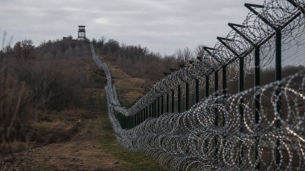 A barbed-wire border fence line running along a quiet wooded ridge with a distant watchtower