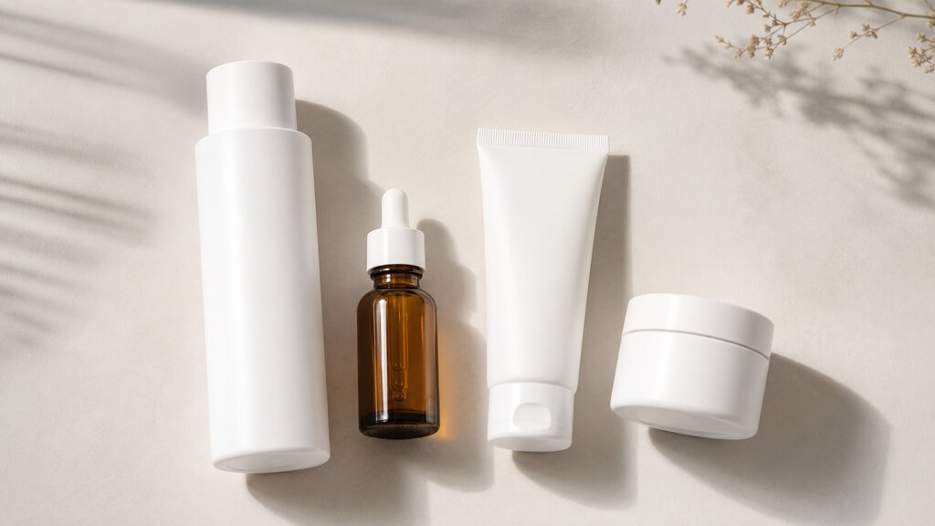 A flat-lay of typical Korean skincare formats on a pale surface — a tall toner bottle, a glass dropper serum, a squeeze sunscreen tube and a small round cream jar, all plain and unlabeled